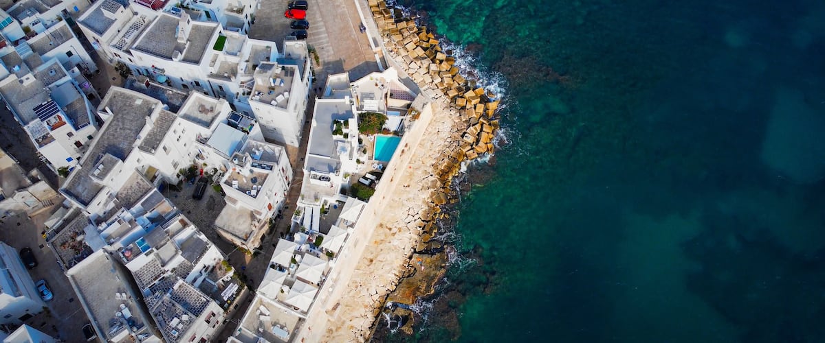 An aerial view of a coastal town with white buildings, a clear blue sea, and a rocky shoreline.