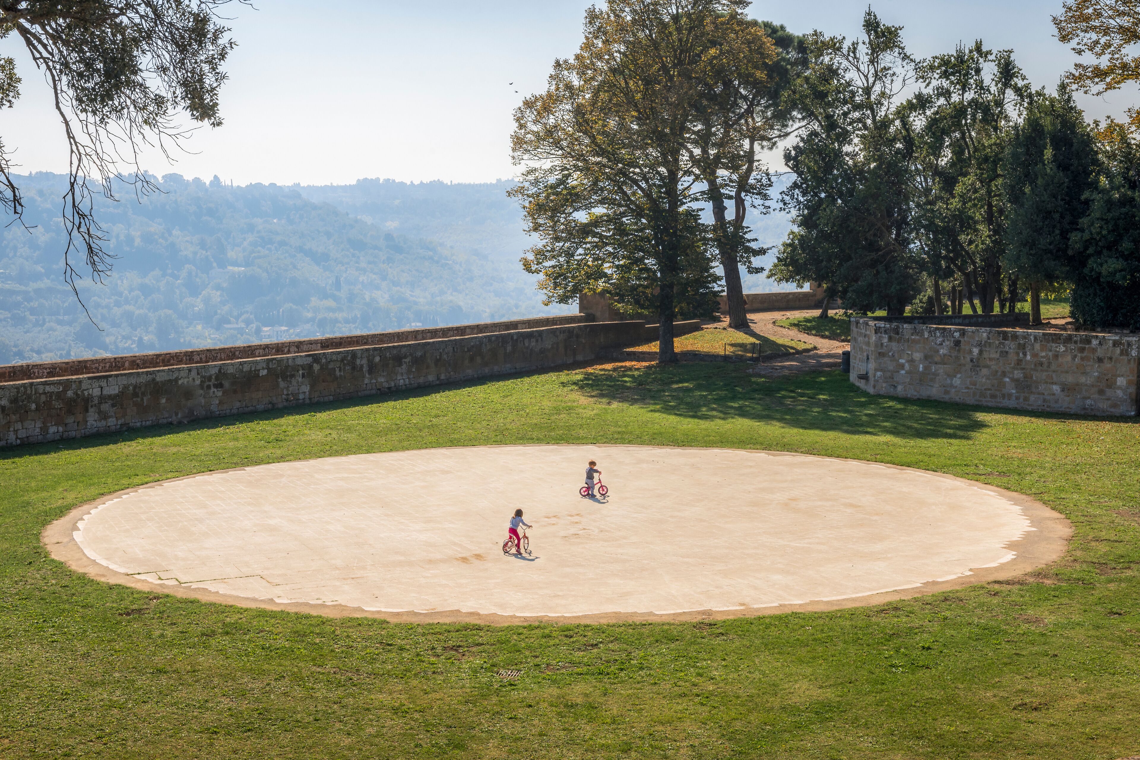 Uma área circular pavimentada com grama, árvores e uma vista distante de uma paisagem montanhosa.