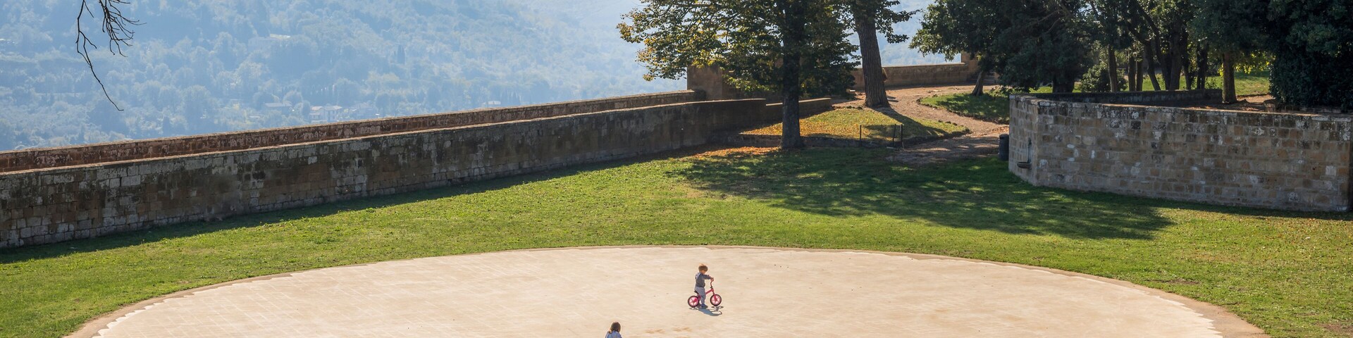 Uma área circular pavimentada com grama, árvores e uma vista distante de uma paisagem montanhosa.