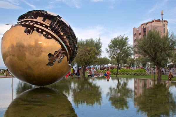 Una grande sfera dorata a forma di scultura in una piazza pubblica, con il suo riflesso nell'acqua, circondata da alberi e un edificio sullo sfondo.