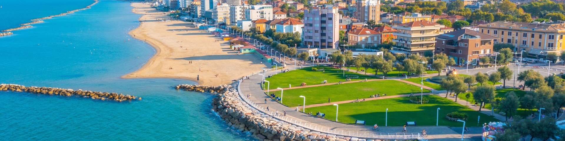 Un paisaje urbano costero con una playa arenosa, un rompeolas rocoso y una mezcla de edificios residenciales y comerciales.