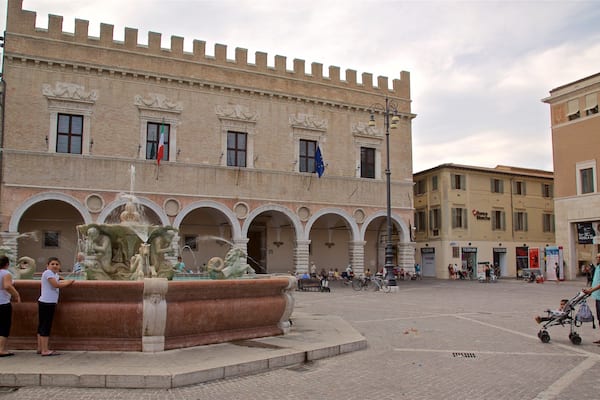 A historic square with a fountain, a castle-like building, and people walking around.