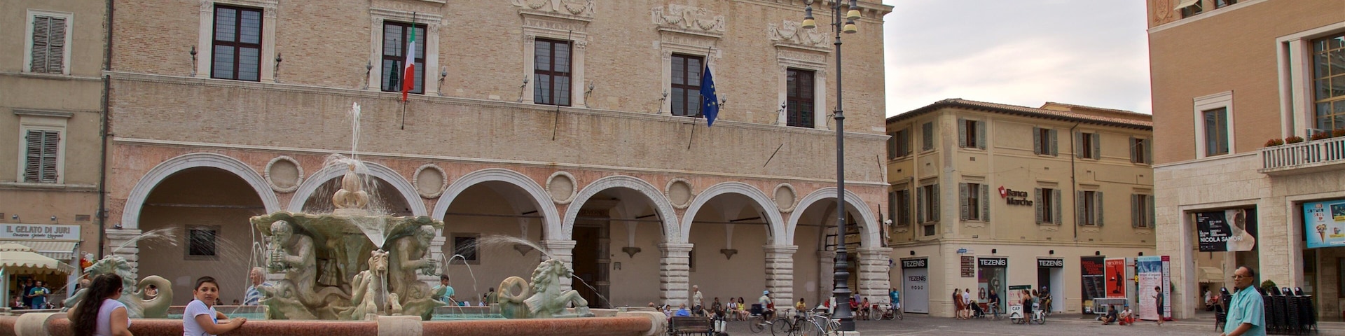Uma praça histórica com uma fonte, um edifício semelhante a um castelo e pessoas caminhando.