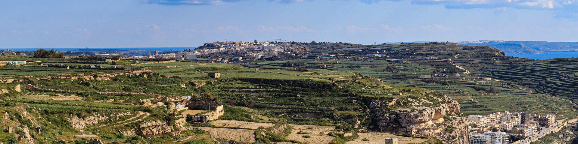 A coastal landscape with terraced fields, ruins, and a distant town.
