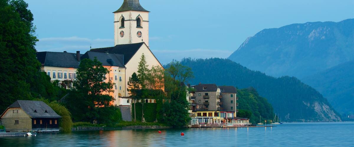 A lakeside town with a church tower, buildings, and mountains in the background.