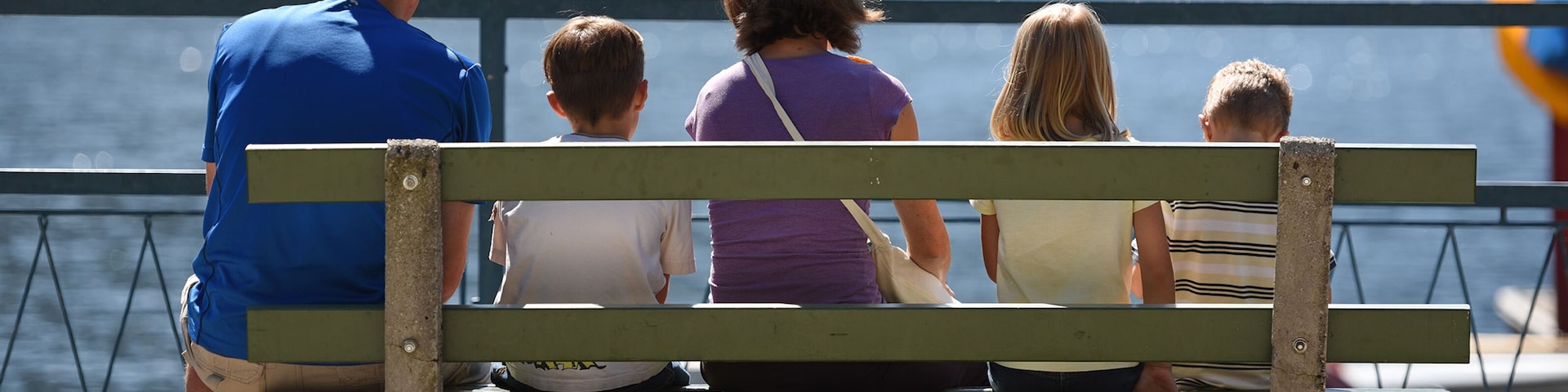 Uma família de cinco sentada em um banco à beira-mar, com guarda-corpo e um corpo de água ao fundo.
