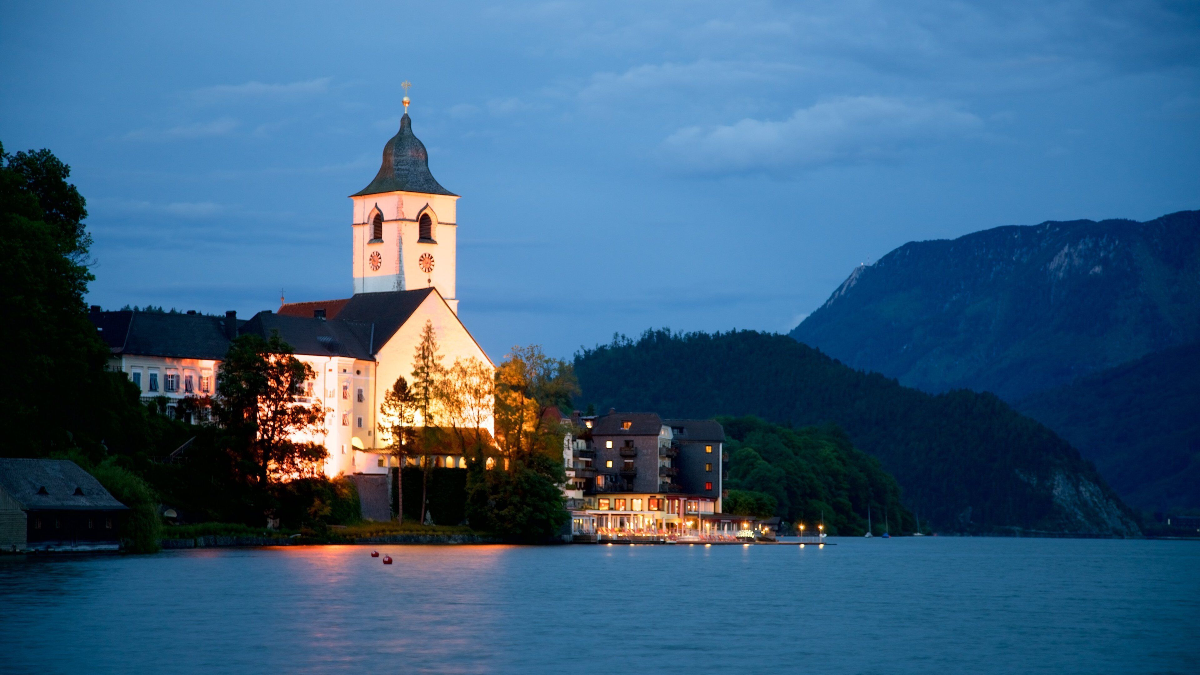 A church with a clock tower by a lake, surrounded by mountains and residential buildings.