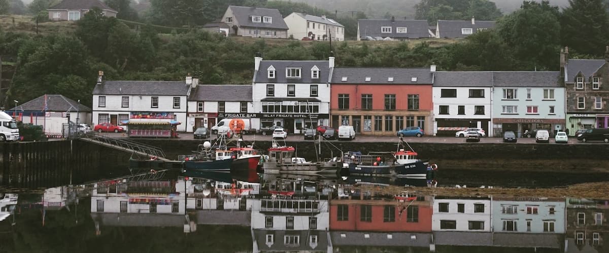 A coastal town with buildings reflecting on calm water, surrounded by fog and greenery.