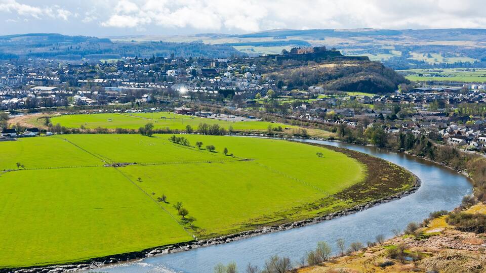 A river winding through a town with green fields and a hill with a castle in the distance.