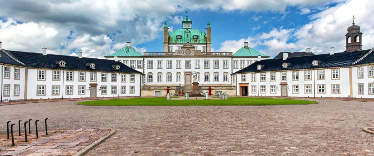 A historic building with a central green dome, symmetrical wings, and a cobblestone courtyard.