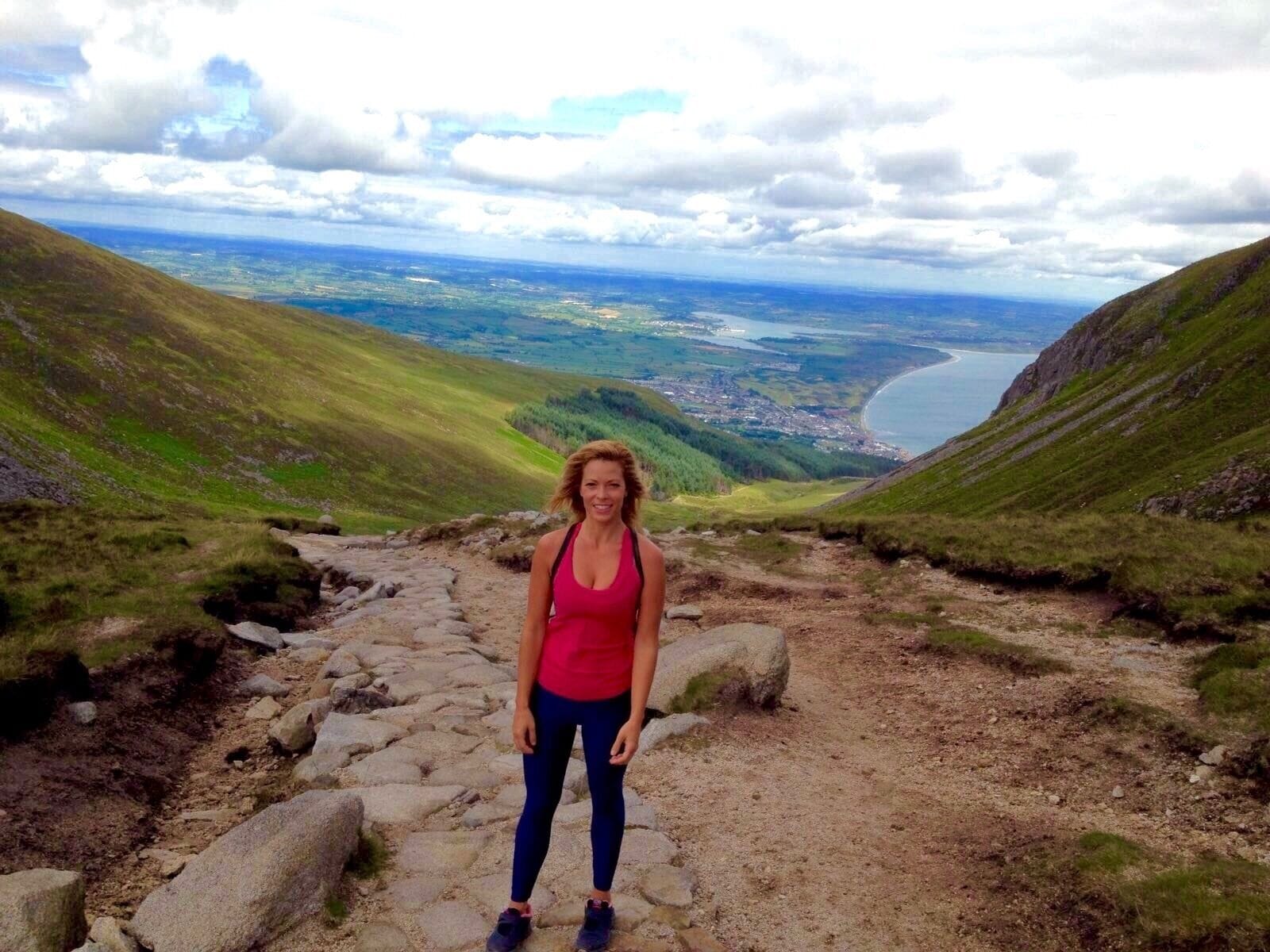 A person standing on a rocky trail with a view of a valley and a body of water in the distance.