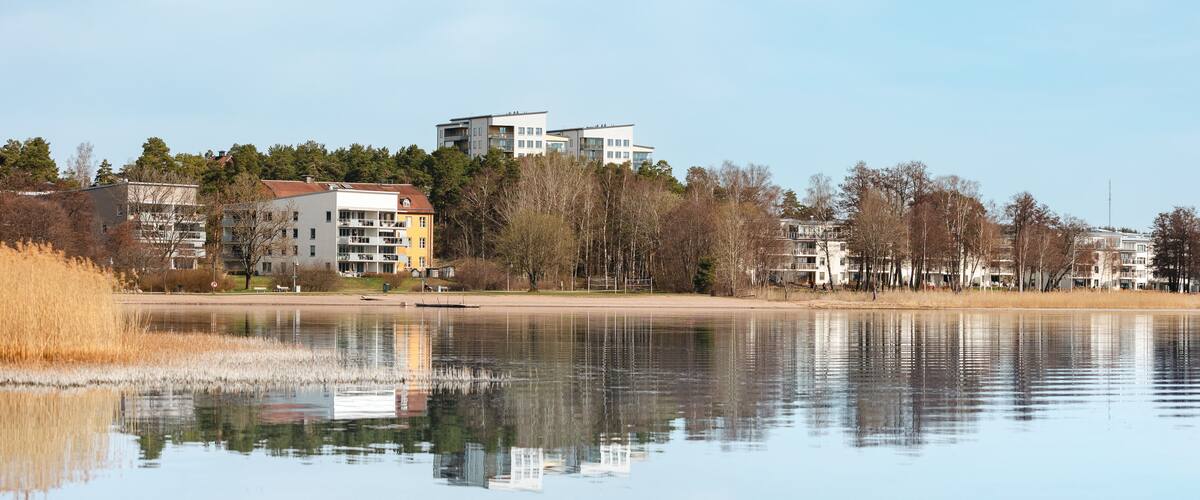 A calm lake with reflections of buildings and trees, surrounded by a mix of residential and natural landscapes.