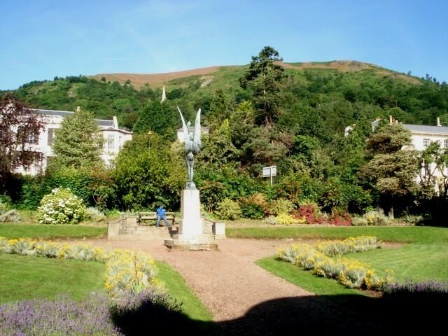 Um jardim com uma estátua, uma colina ao fundo e casas.