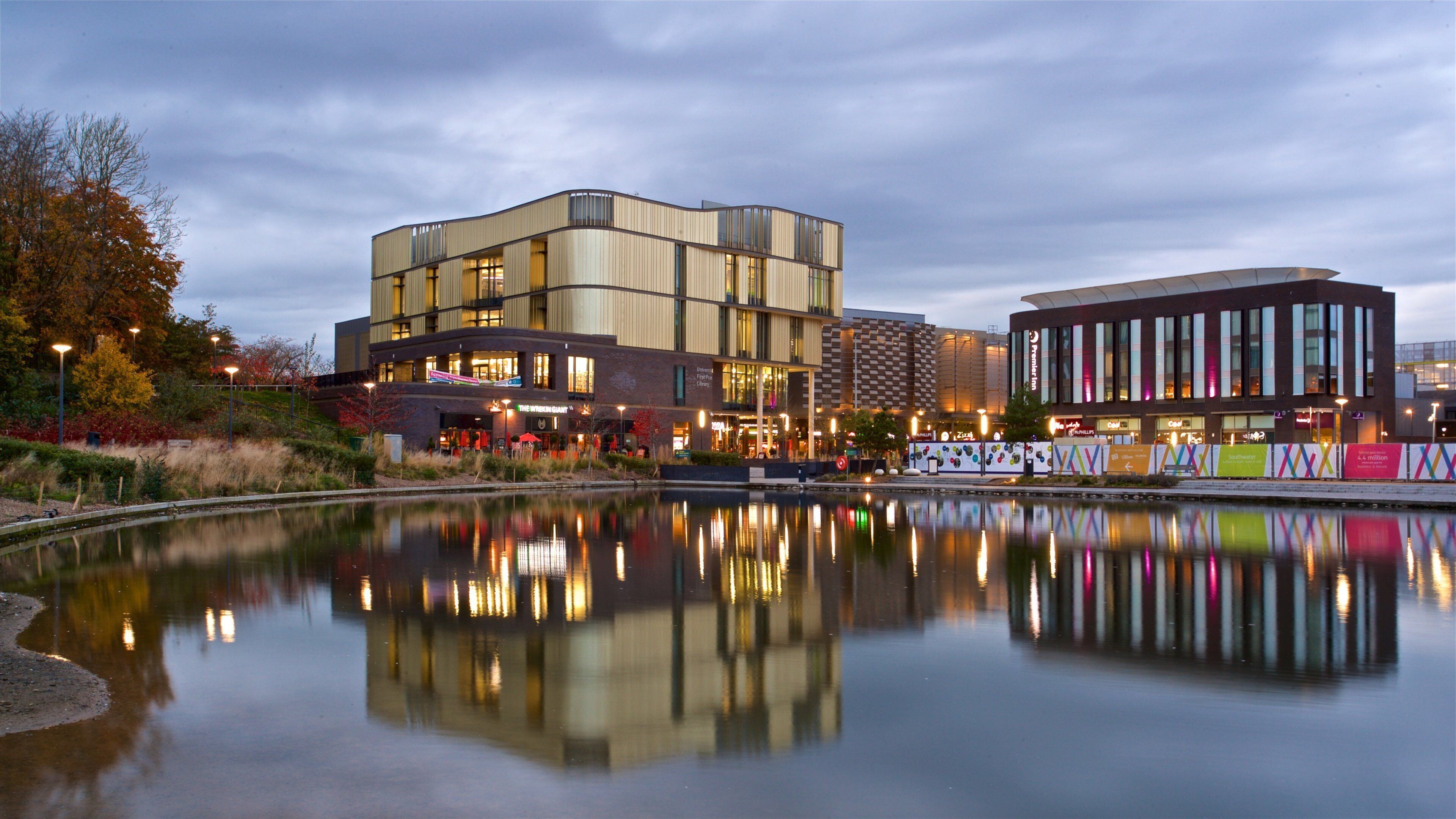 A modern building complex with a reflection in a calm water body, surrounded by trees and urban structures.