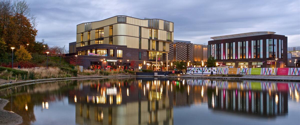 A modern building complex with a reflection in a calm water body, surrounded by trees and urban structures.