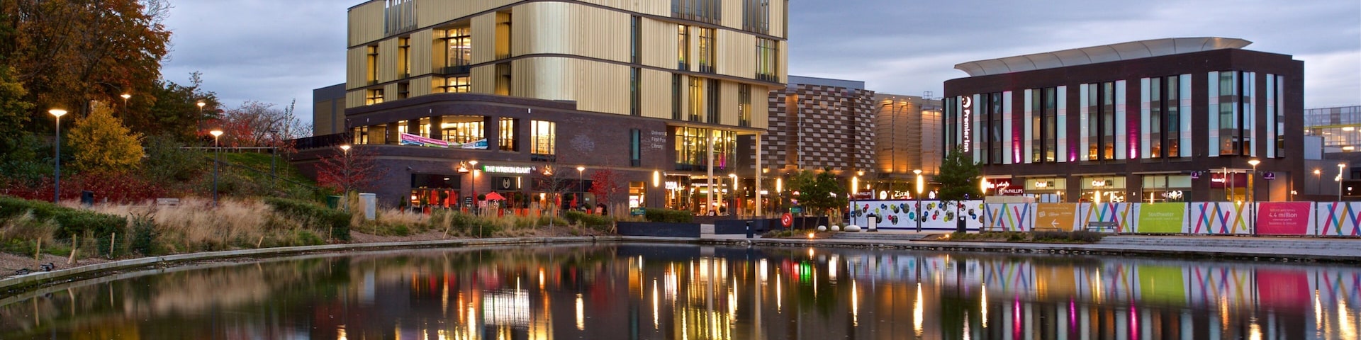 A modern building complex with a reflection in a calm water body, surrounded by trees and urban structures.