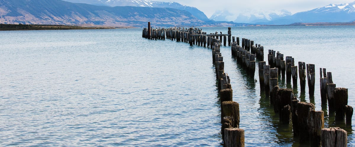 A wooden pier extending into a calm lake with snow-capped mountains in the background.