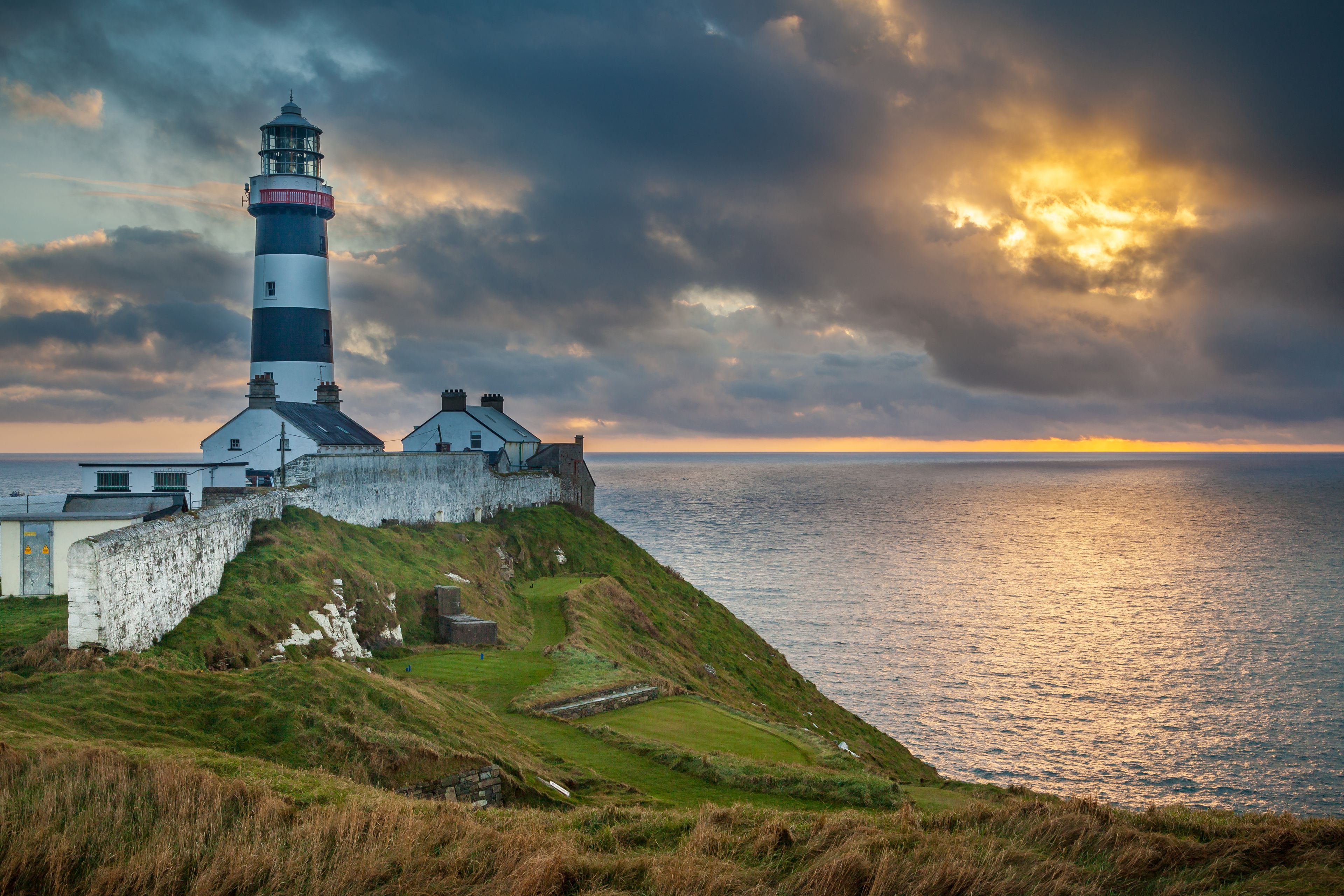 A lighthouse on a grassy cliff overlooking the sea, with a dramatic sky and sunset.