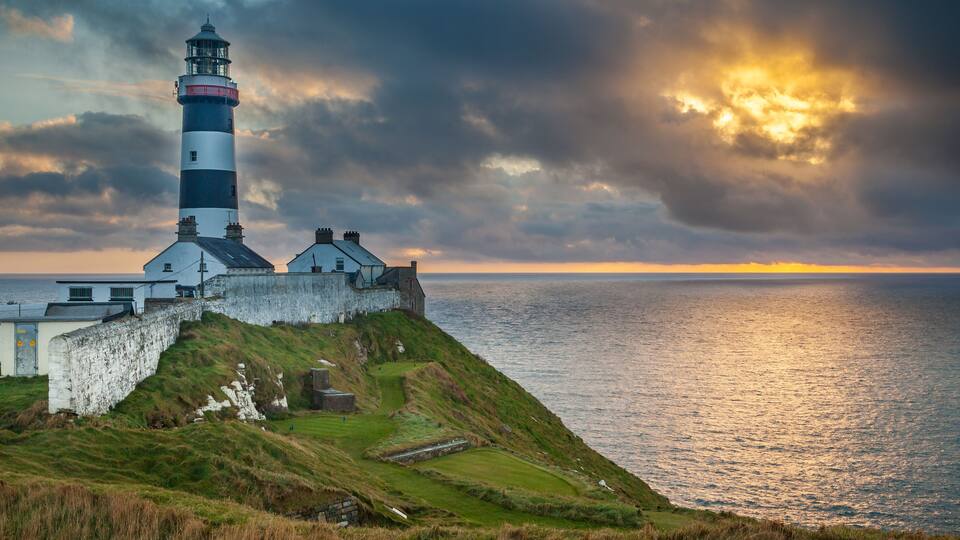 A lighthouse on a grassy cliff overlooking the sea, with a dramatic sky and sunset.