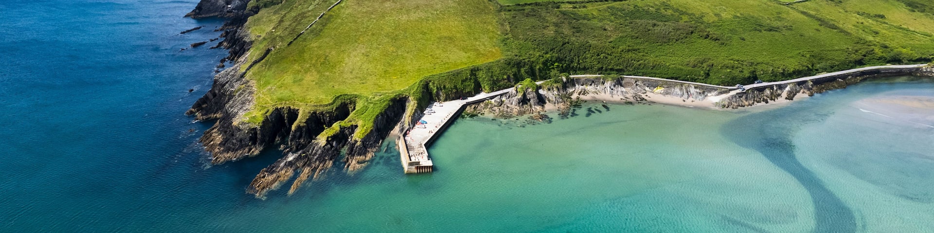 A coastal landscape with a clear sky, a rocky coastline, a green field, and a calm sea.