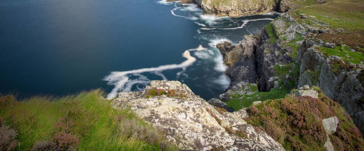 A coastal cliff with waves crashing against it, a grassy hillside, and a distant rocky outcrop.