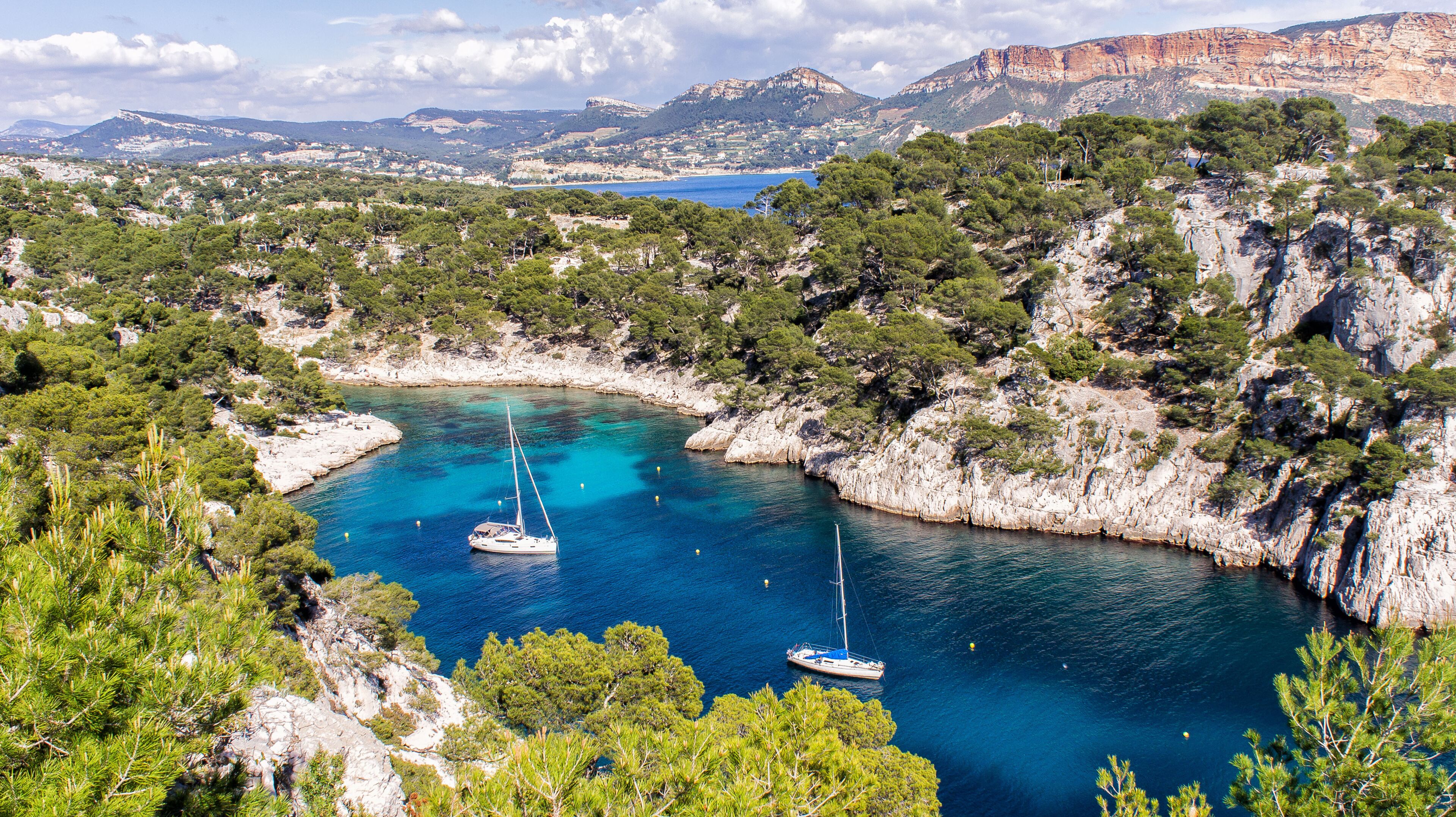A coastal bay with clear turquoise waters, surrounded by rocky cliffs and dense green pine trees. Two sailboats are anchored in the bay.
