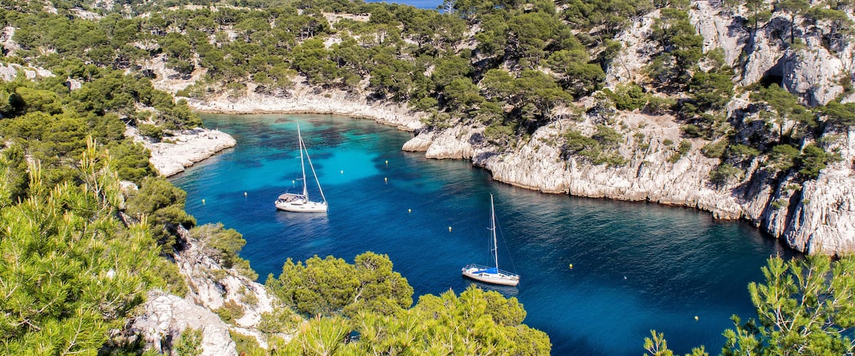 A coastal bay with clear turquoise waters, surrounded by rocky cliffs and dense green pine trees. Two sailboats are anchored in the bay.