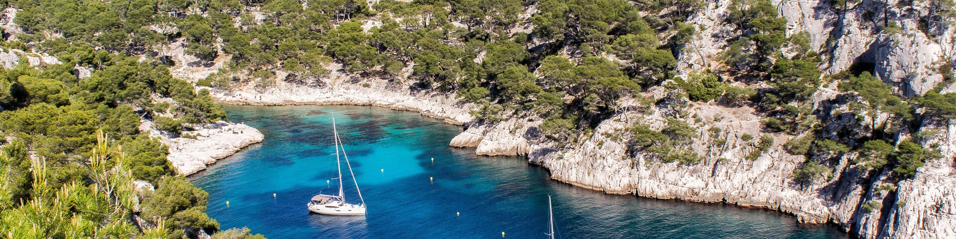 A coastal bay with clear turquoise waters, surrounded by rocky cliffs and dense green pine trees. Two sailboats are anchored in the bay.