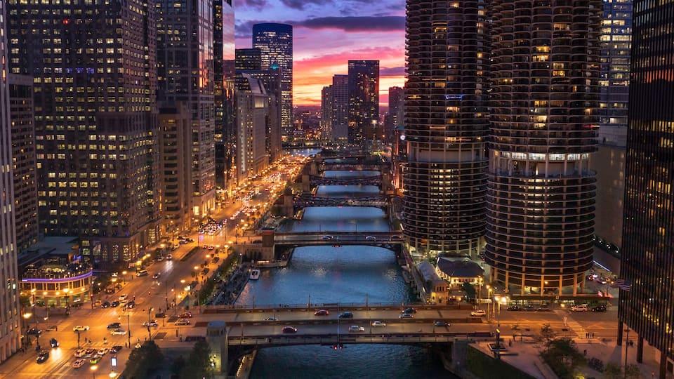 A cityscape with tall buildings, a river, and a bridge at dusk.