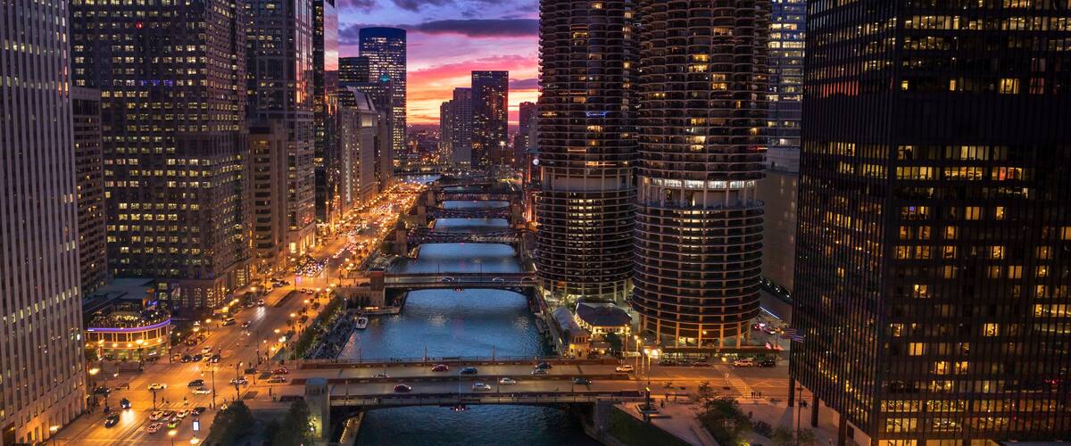 A cityscape with tall buildings, a river, and a bridge at dusk.