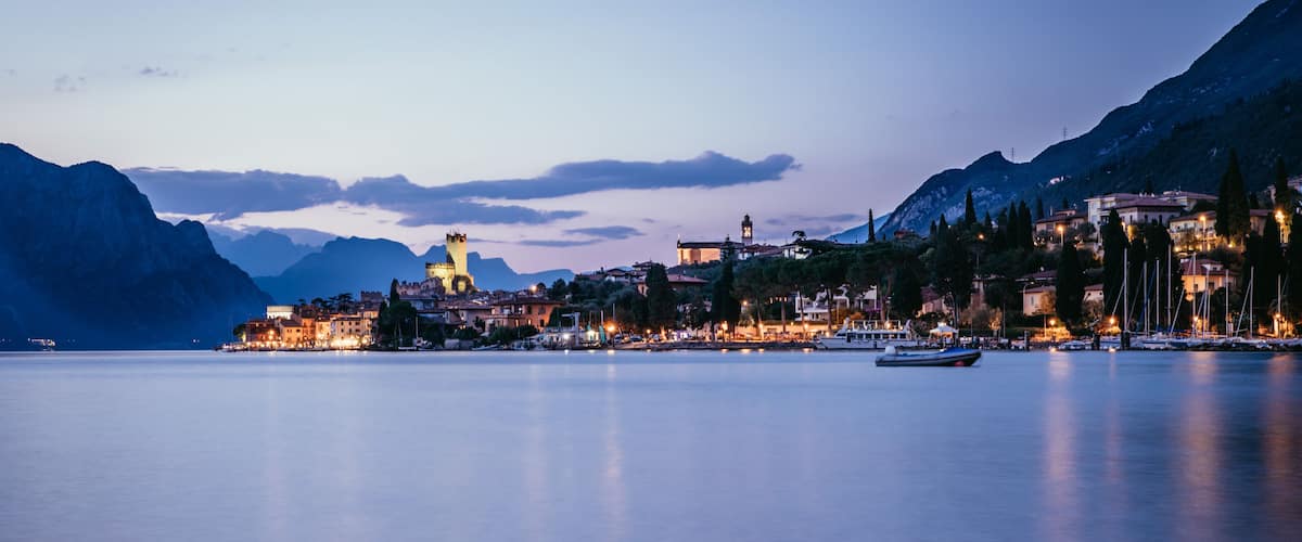 A calm lake with a town on the shore, mountains in the background, and boats in the water.