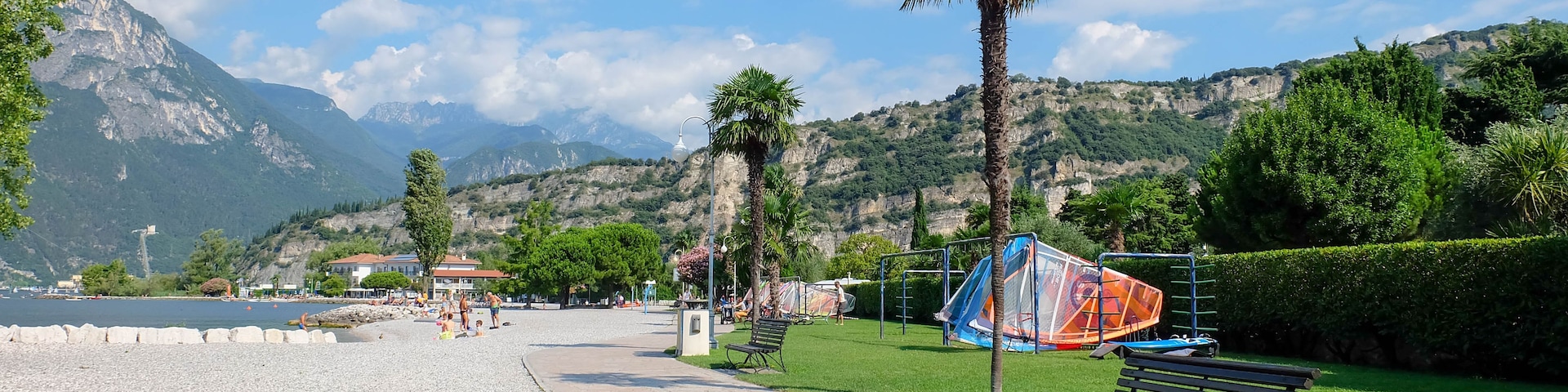 Um parque à beira do lago com um caminho de pedrinhas, bancos, palmeiras e um veleiro.