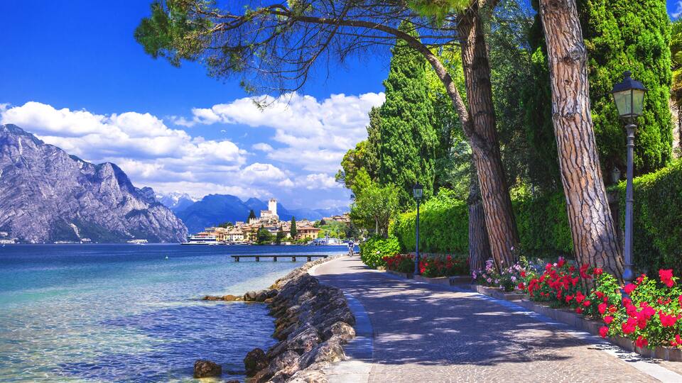 A coastal walkway with clear water, rocky shore, and mountains in the background.