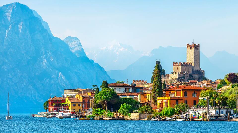 A coastal town with colorful buildings, a castle, and mountains in the background.