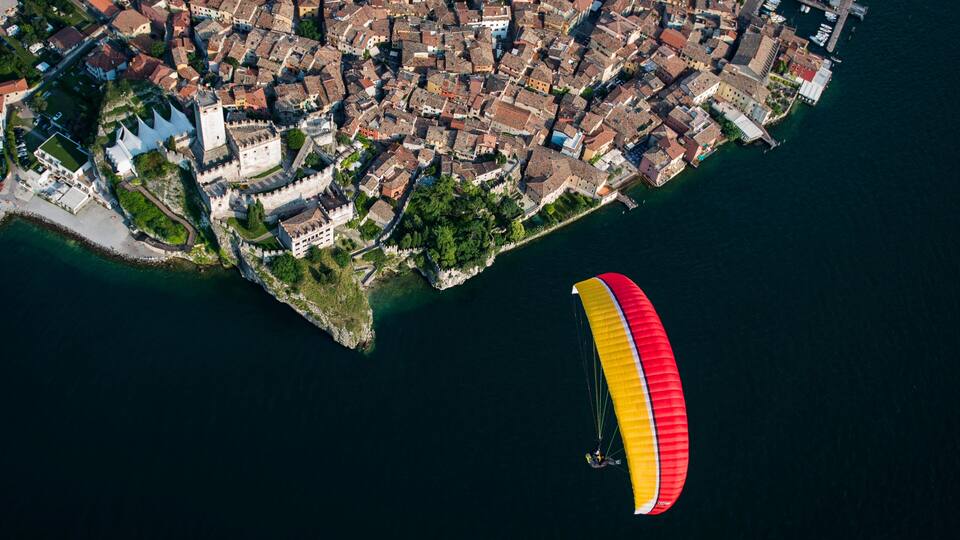 An aerial view of a coastal town with dense historic buildings, a prominent church, and a person paragliding nearby.
