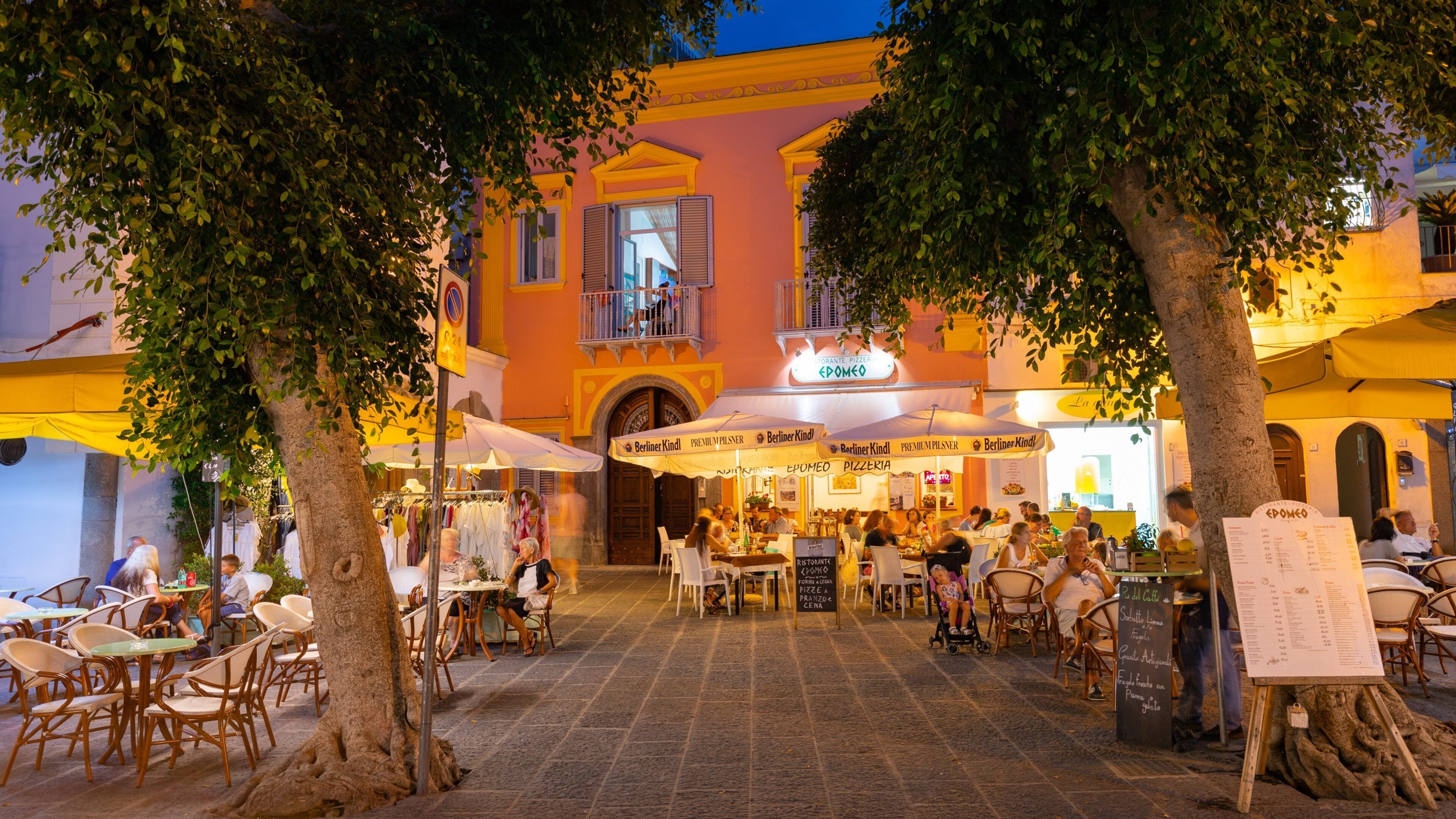 An outdoor cafe with tables and chairs, surrounded by trees and buildings with yellow and orange facades.