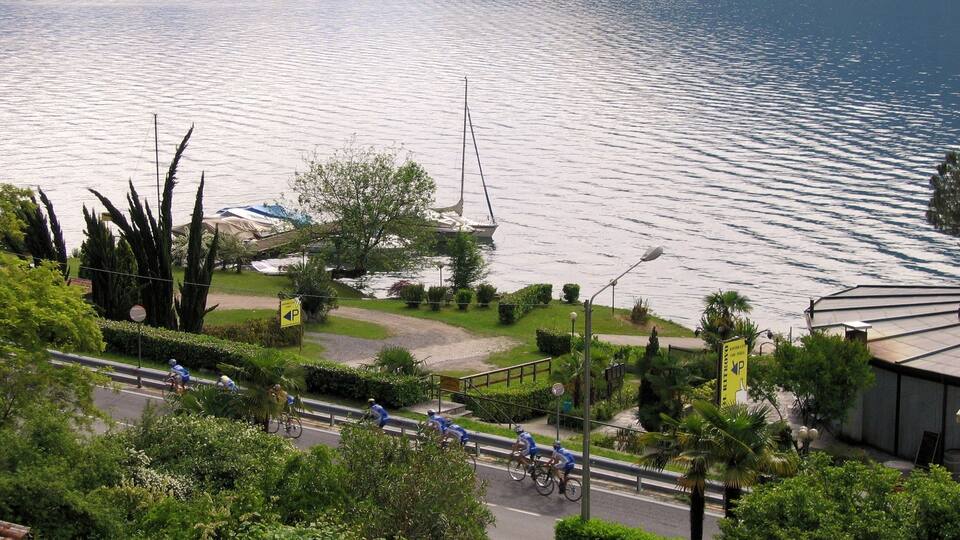 A lake with a sailboat, cyclists on a path, and greenery.