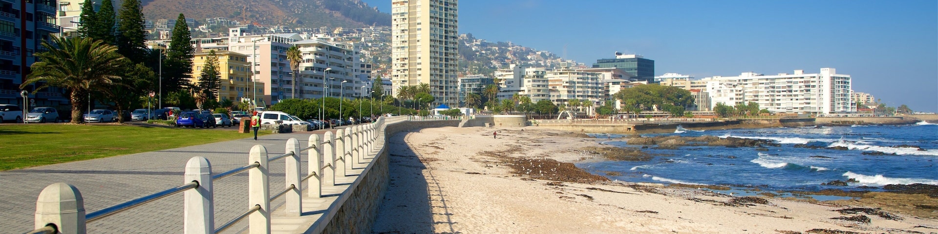 Un paysage urbain côtier, avec une montagne imposante en arrière-plan, une plage de sable et une rangée de bâtiments modernes le long de la promenade.