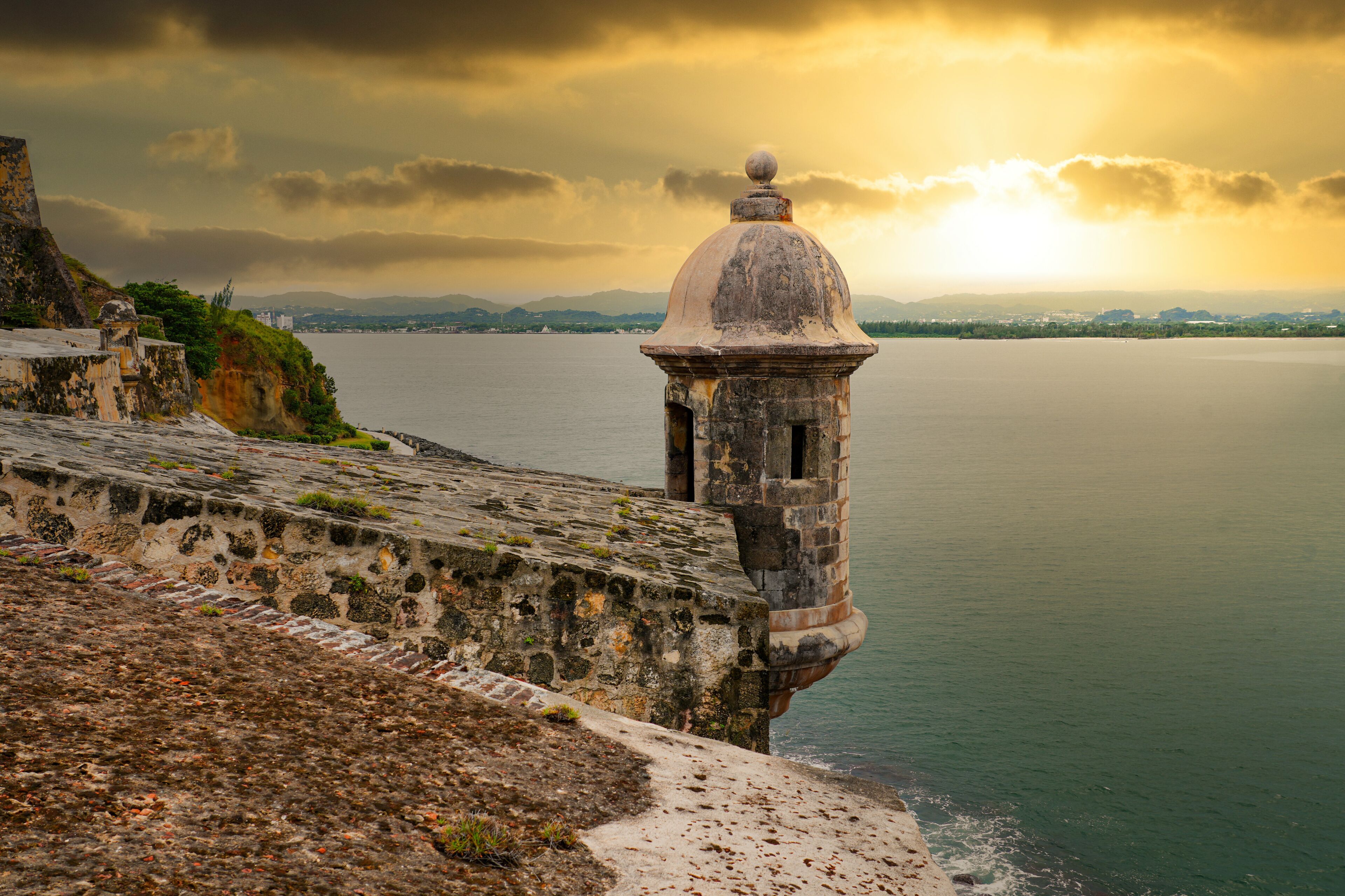 A coastal fortification with a domed tower overlooking a calm sea, with mountains in the background under a cloudy sky.