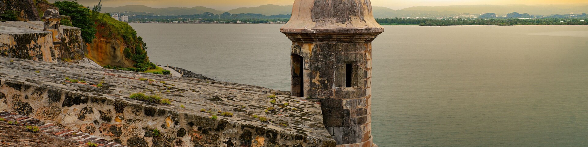 A coastal fortification with a domed tower overlooking a calm sea, with mountains in the background under a cloudy sky.