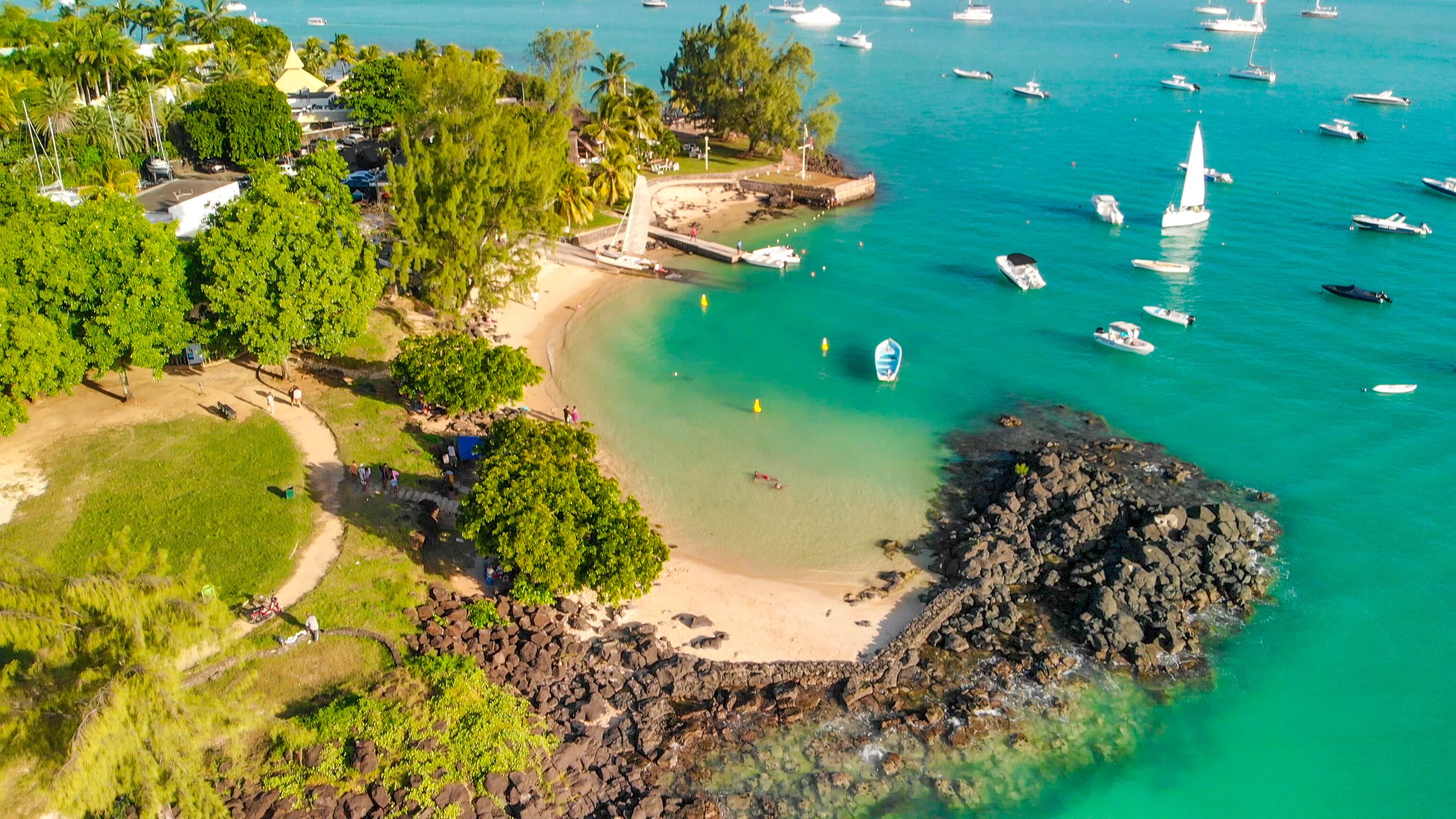 An aerial view of a coastal area with a sandy beach, rocky shoreline, clear turquoise waters, and numerous boats.
