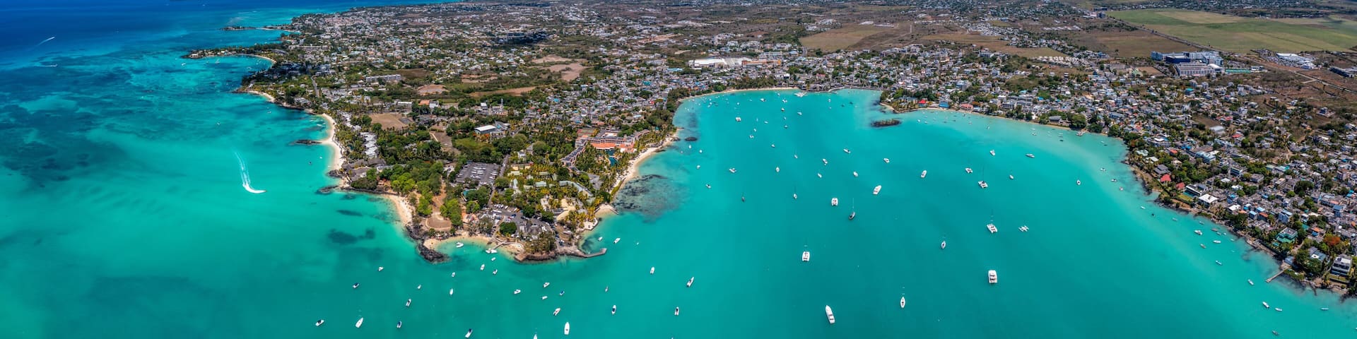 An aerial view of a coastal town with clear turquoise waters, a densely populated area, and a large bay with numerous boats.