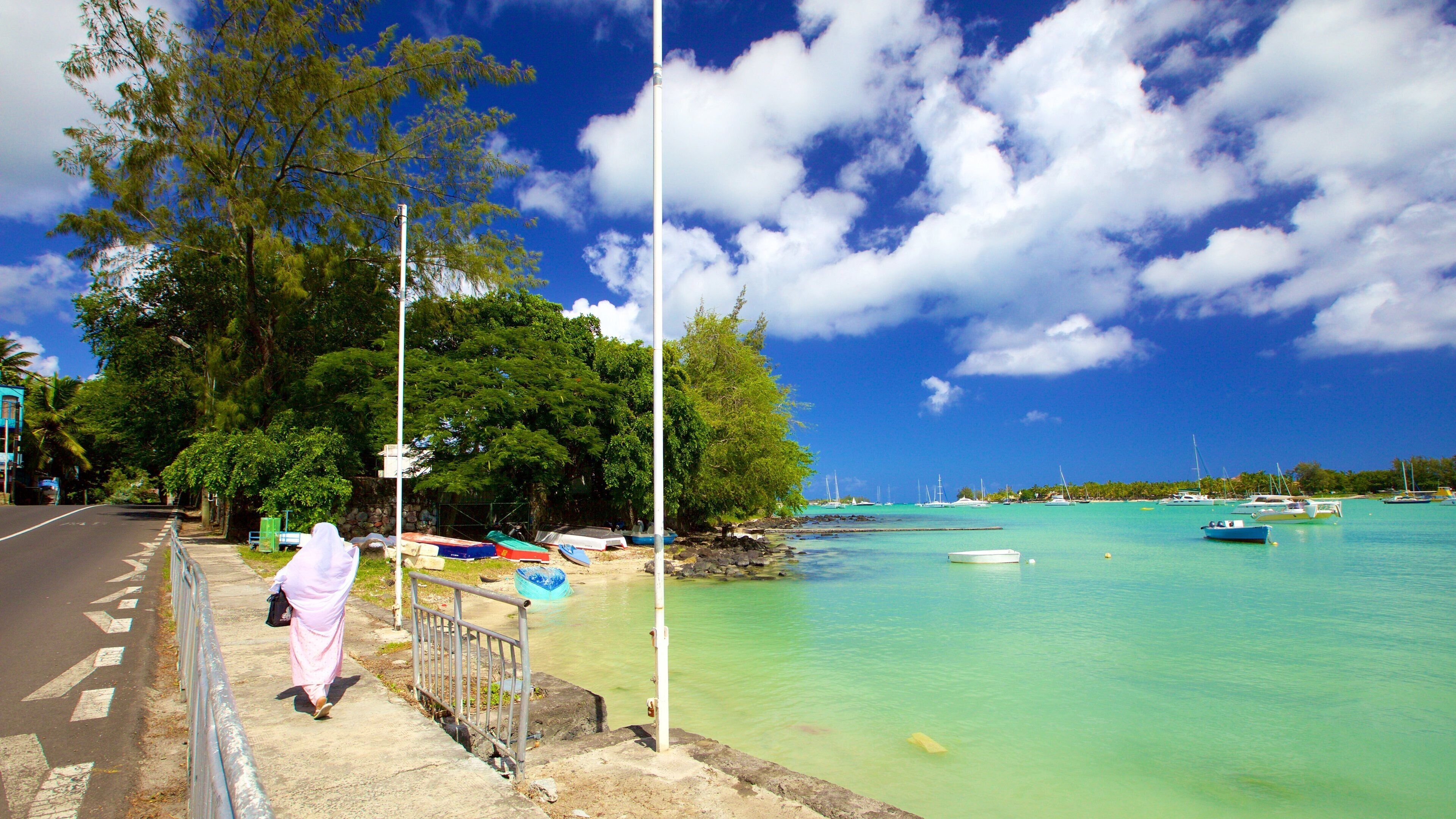 A coastal road with a pedestrian walkway, clear turquoise waters, boats, and lush greenery.
