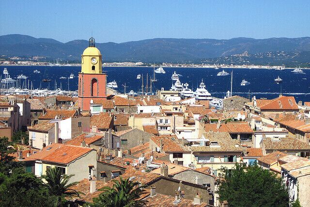 Un pueblo costero con una imponente torre del reloj amarilla, edificios con tejados de tejas rojas y un puerto deportivo lleno de barcos.
