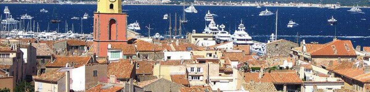 A coastal town with a prominent yellow clock tower, terracotta-roofed buildings, and a marina with boats.