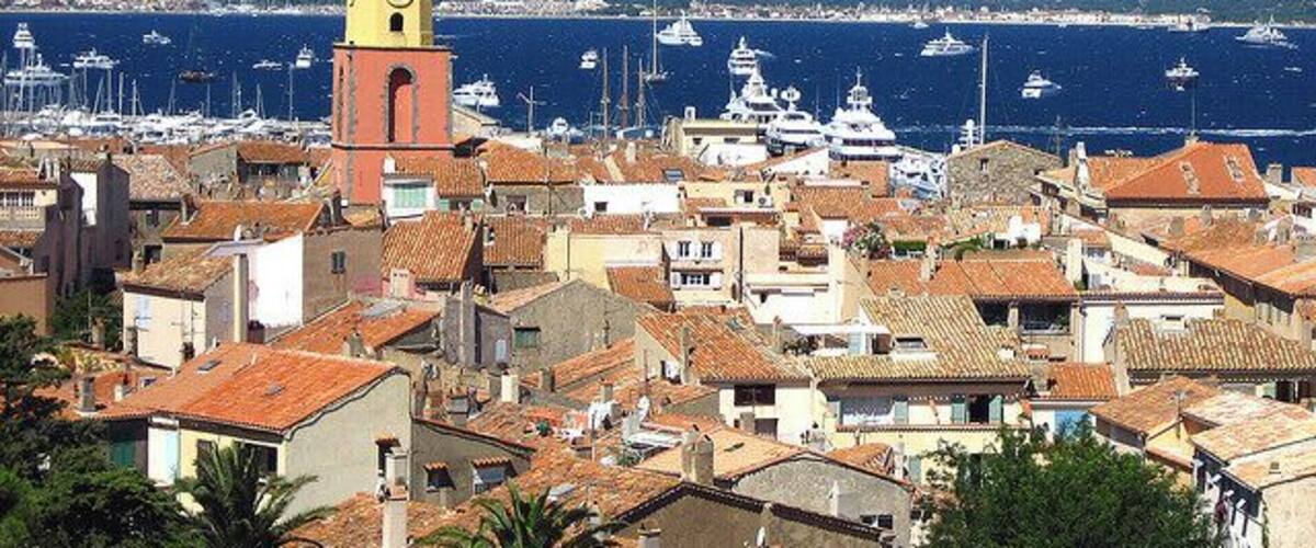 A coastal town with a prominent yellow clock tower, terracotta-roofed buildings, and a marina with boats.