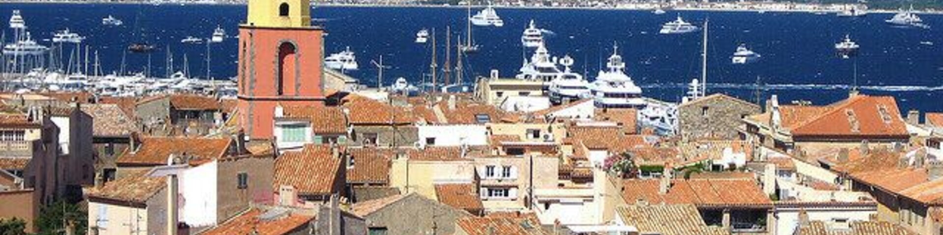 Un pueblo costero con una imponente torre del reloj amarilla, edificios con tejados de tejas rojas y un puerto deportivo lleno de barcos.