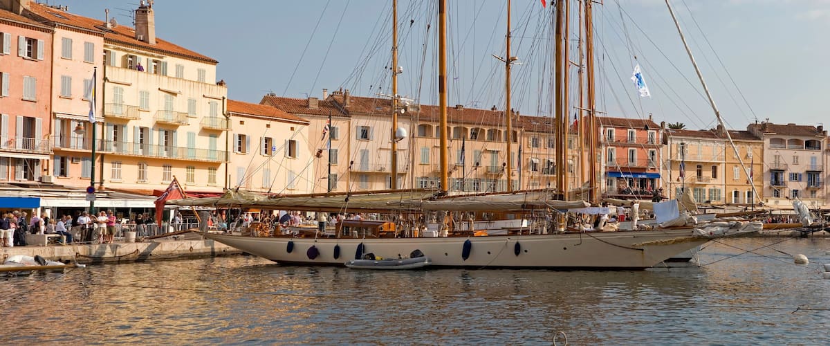 A historic waterfront with tall sailing ships docked, surrounded by colorful buildings and a crowd of people.