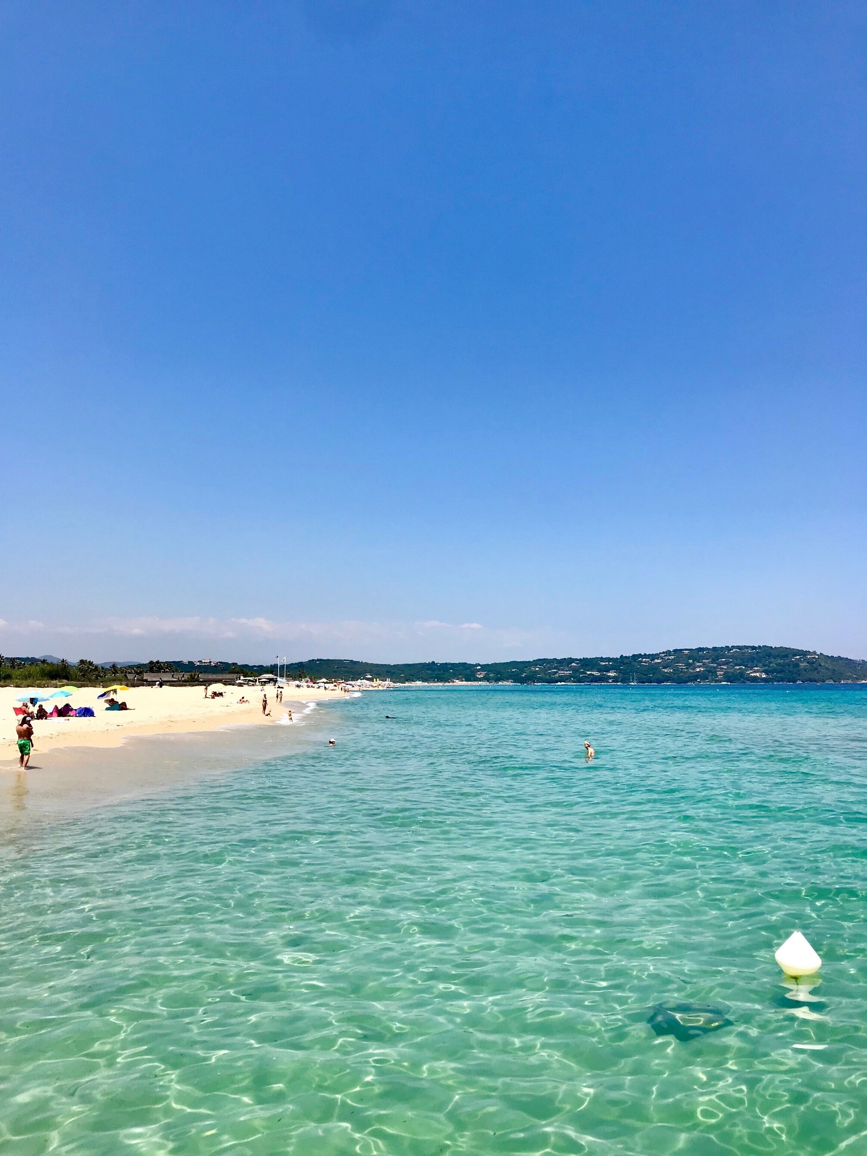 Una playa con agua turquesa cristalina, gente tomando el sol y una colina distante con edificios.
