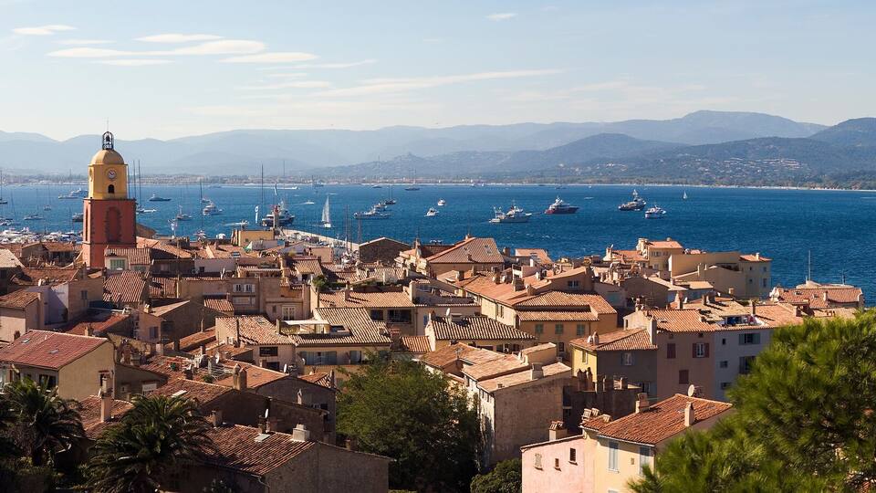 A coastal town with a prominent tower, surrounded by mountains and a clear blue sea with sailboats.